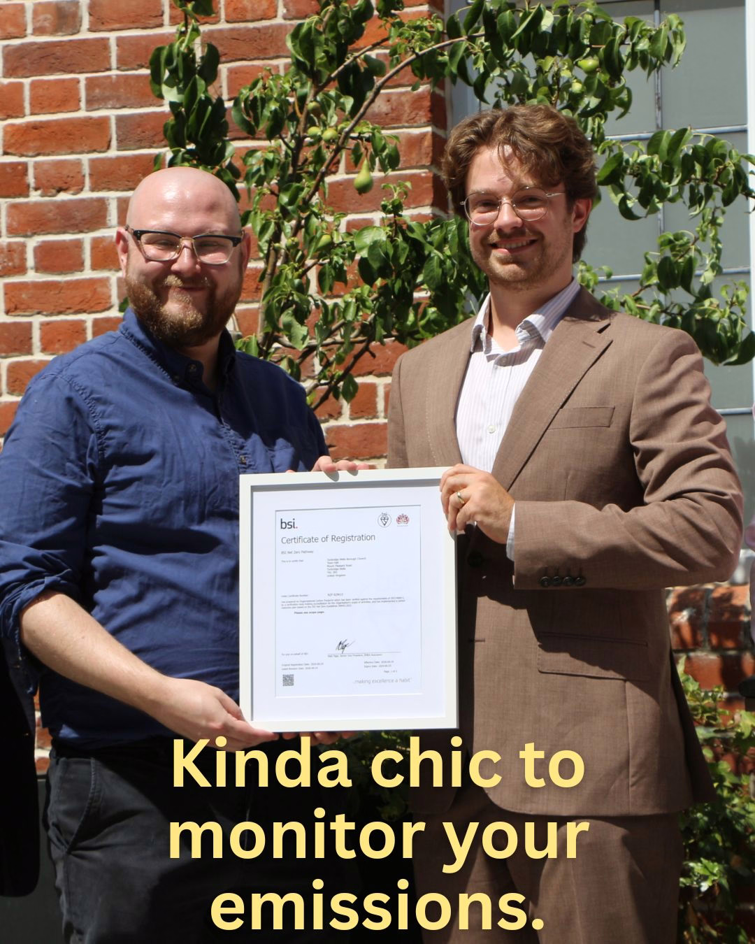 Two smiling men (one in a brown suit, one in a blue shirt) hold a framed BSI Certificate of Registration outdoors against a brick wall; yellow overlay text reads "Kinda chic to monitor your emissions," celebrating certification for emissions monitoring.