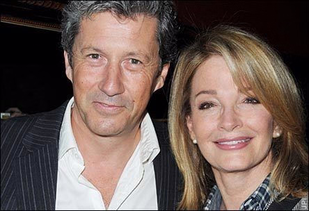 A candid behind-the-scenes photo from around 2012 featuring Deidre Hall and Charles Shaughnessy, known for their roles as Marlena Evans and Shane Donovan on Days of Our Lives. Deidre wears a plaid shirt layered under a tweed blazer, with her blonde hair styled straight and shoulder-length. Charles wears a dark pinstripe jacket over an unbuttoned white shirt. Both are smiling warmly and looking directly at the camera. The background is dark, suggesting an indoor setting with subdued lighting. The image evokes a sense of warmth, legacy, and quiet drama, capturing two soap opera icons during a storyline involving safe houses and high-stakes intrigue.
