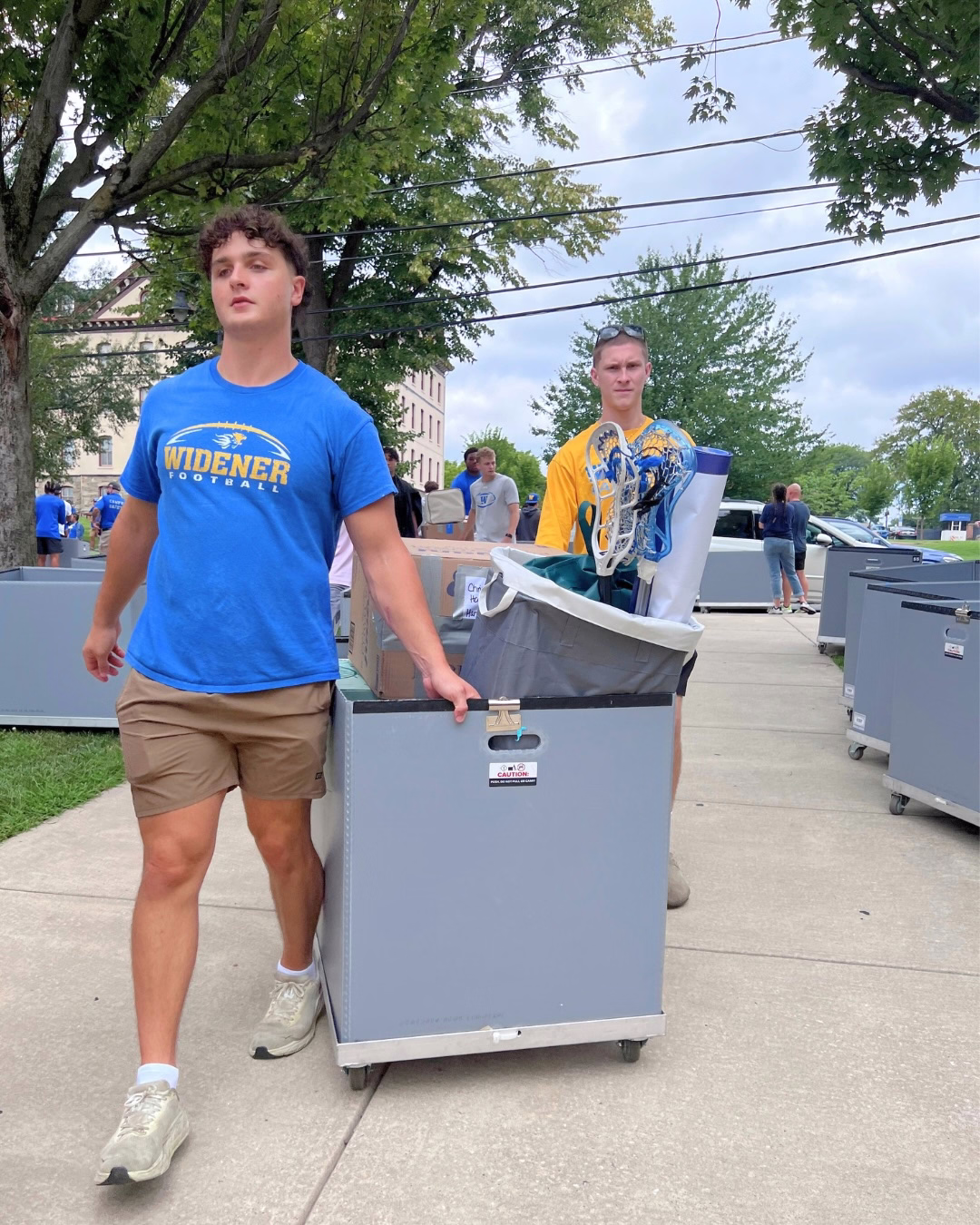 A student in Widener football shirt pulls a bin full of dorm items plus two lacrosse sticks, as a student follows behind