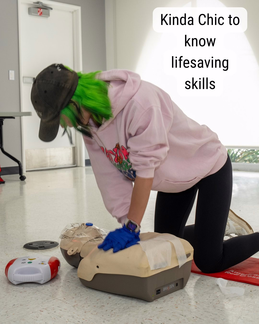 A person with green hair, wearing a hoodie and cap, practices CPR on a mannequin. Nearby is a defibrillator device. A caption reads, "Kinda Chic to know lifesaving skills." The setting appears to be an indoor training space.