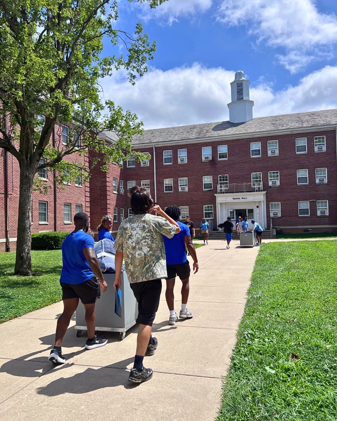 Students push a cart or walk alongside it as they walk down the pathway to Hanna Hall, a dorm building with a cupola on top
