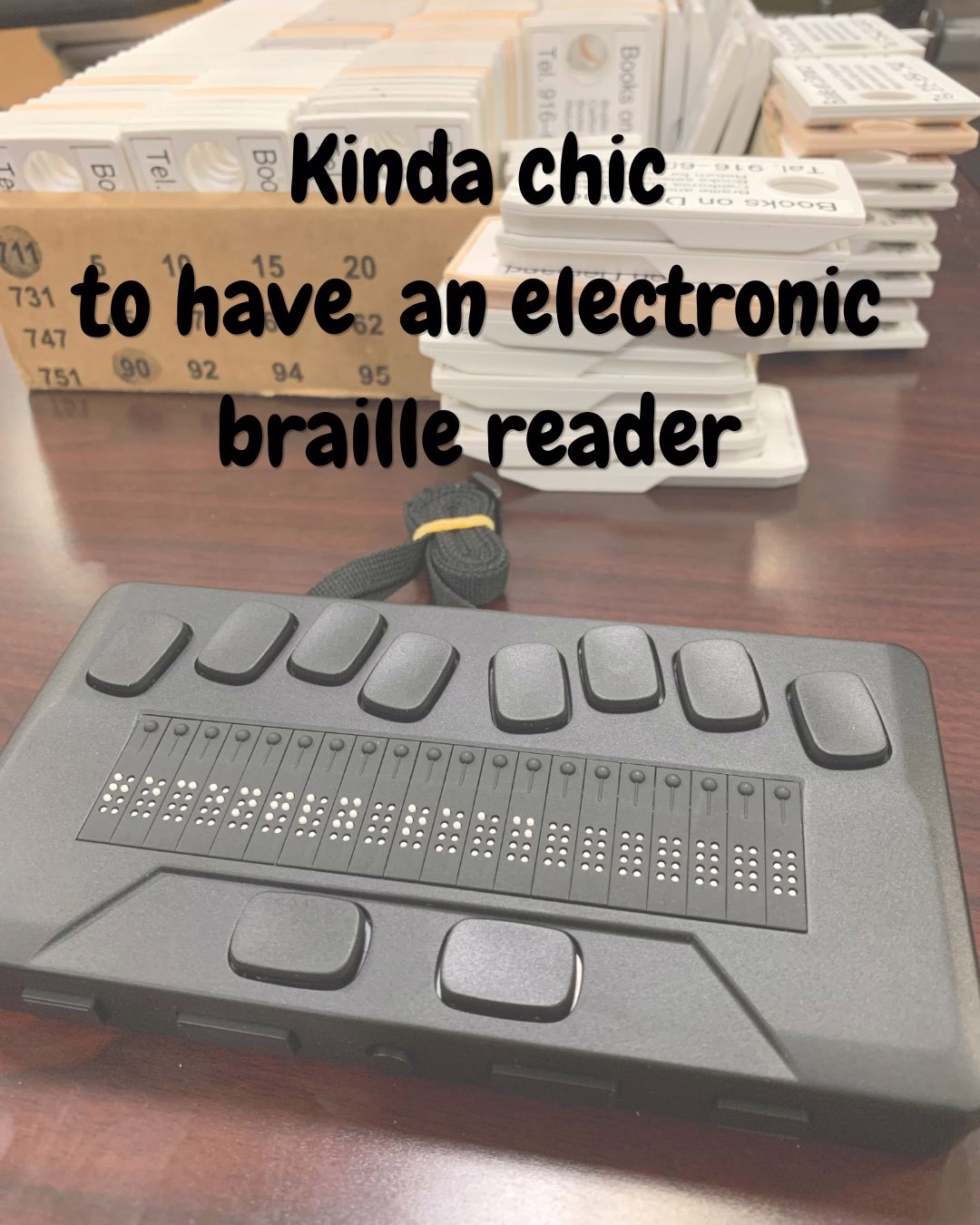 a black electronic braille reader that has 10 large buttons and in the center smaller braille buttons, on a dark wood desk with book flashdrives next to it. black chewy font text overlaid reads "kinda chic to have an electronic braille reader."