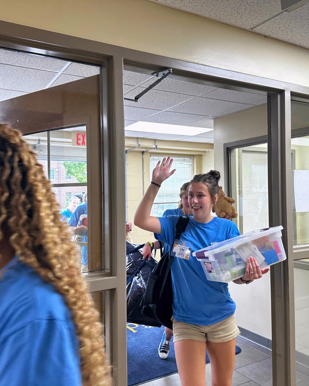 A student holds a bin of dorm items and carries a bag on her shoulder as she enters a dorm building door and waves