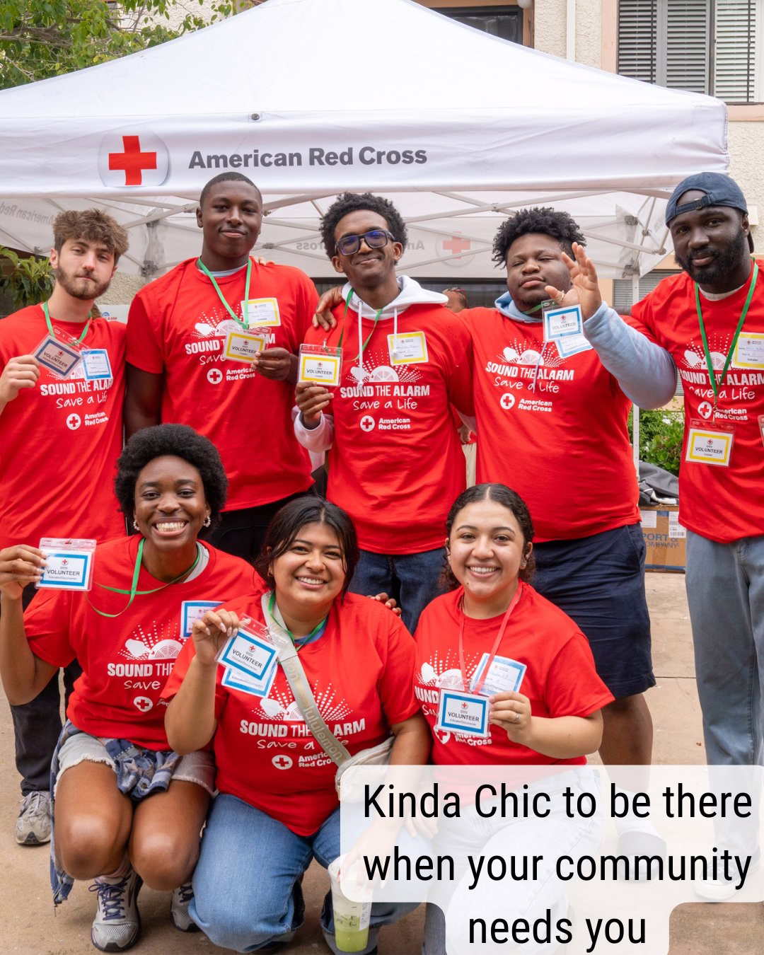 A group of eight people wearing red "Sound the Alarm" T-shirts pose happily under an American Red Cross tent. They hold name badges and stand together outdoors. A message at the bottom reads, "Kinda Chic to be there when your community needs you.