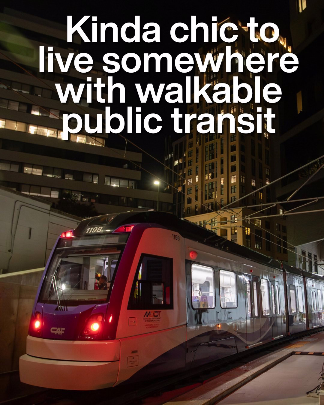 Modern light rail train stopped at a city station at night with illuminated buildings in the background.