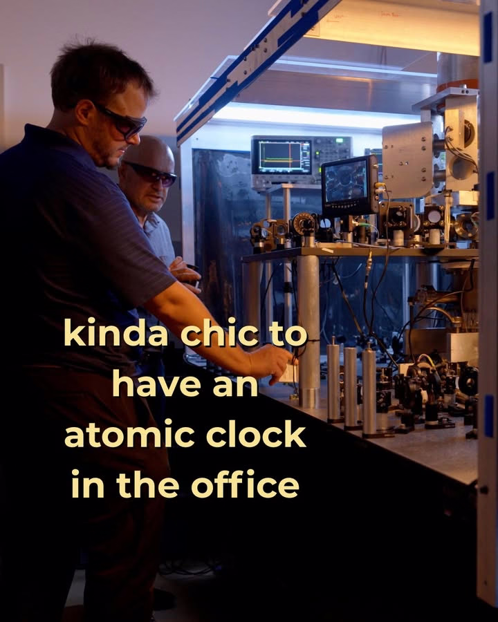Two individuals are observing an atomic clock setup in an office environment. The ambient lighting is dim, highlighting the machinery and screens. Text overlay reads, "kinda chic to have an atomic clock in the office."