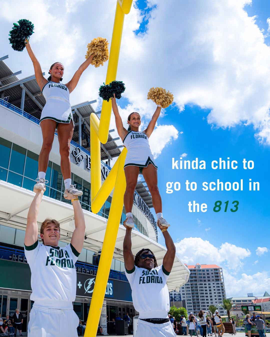 USF cheerleaders performing a stunt outside a Benchmark Arena. Two individuals are holding up two others who are each standing and holding pom-poms. The stadium and a cityscape are visible in the background. Text reads, "kinda chic to go to school in the 813."
