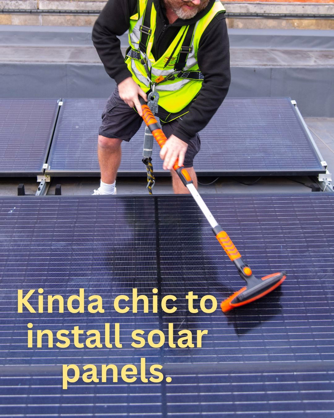 A technician in a high-visibility vest and safety harness scrubs rooftop solar panels with an orange brush; yellow overlay text reads, "Kinda chic to install solar panels."