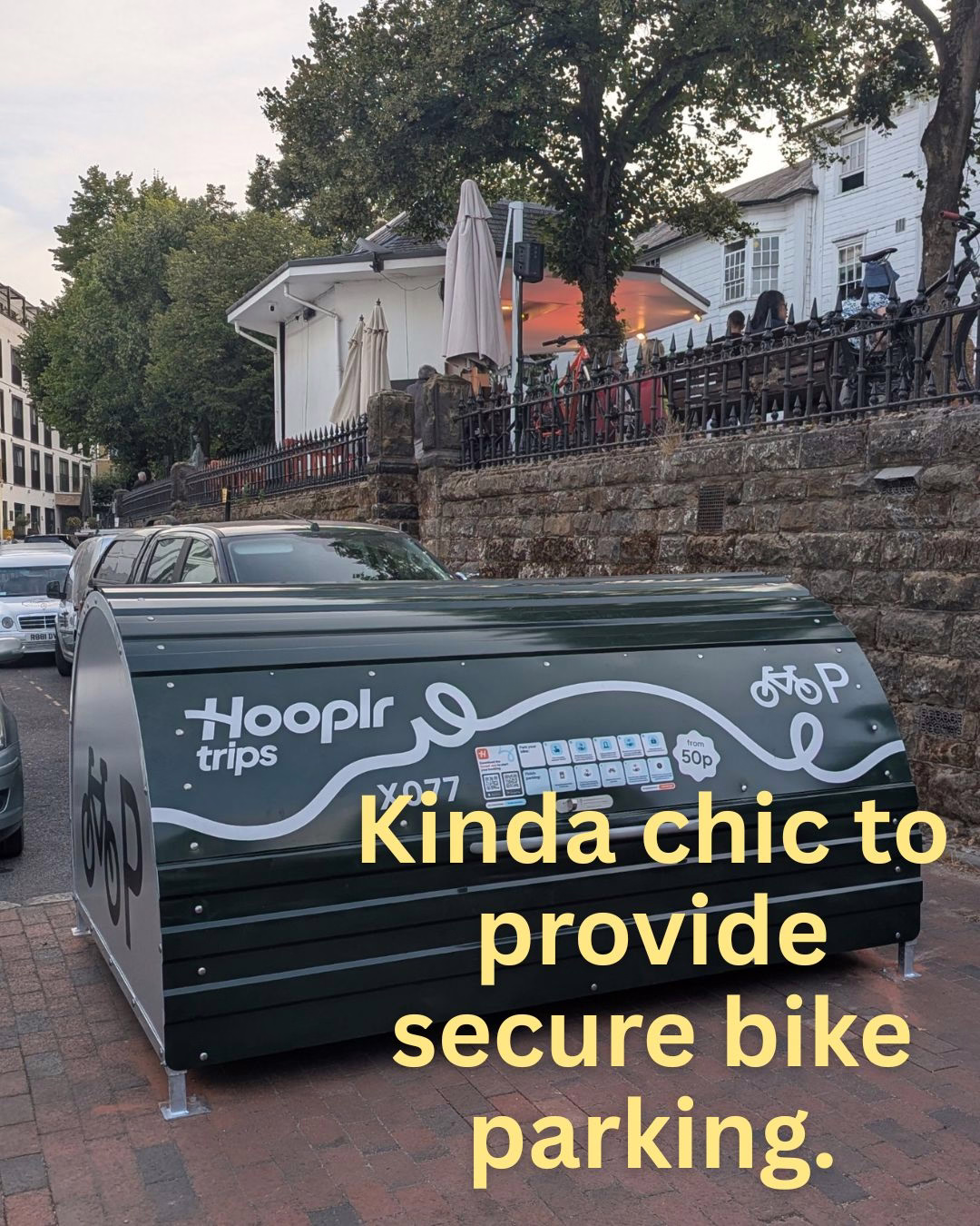 Covered, secure bike storage unit labeled "Hooplr trips" on a city sidewalk beside parked cars and a raised café terrace, with overlaid yellow text reading "Kinda chic to provide secure bike parking."