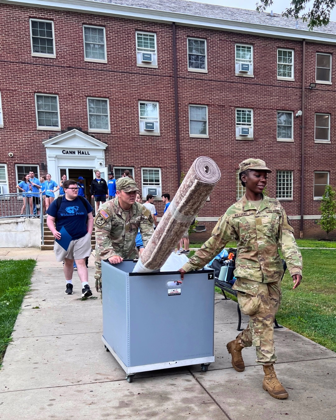Two R-O-T-C cadets in Army fatigues push a cart with a large rug sticking out of it as a student follows behind them