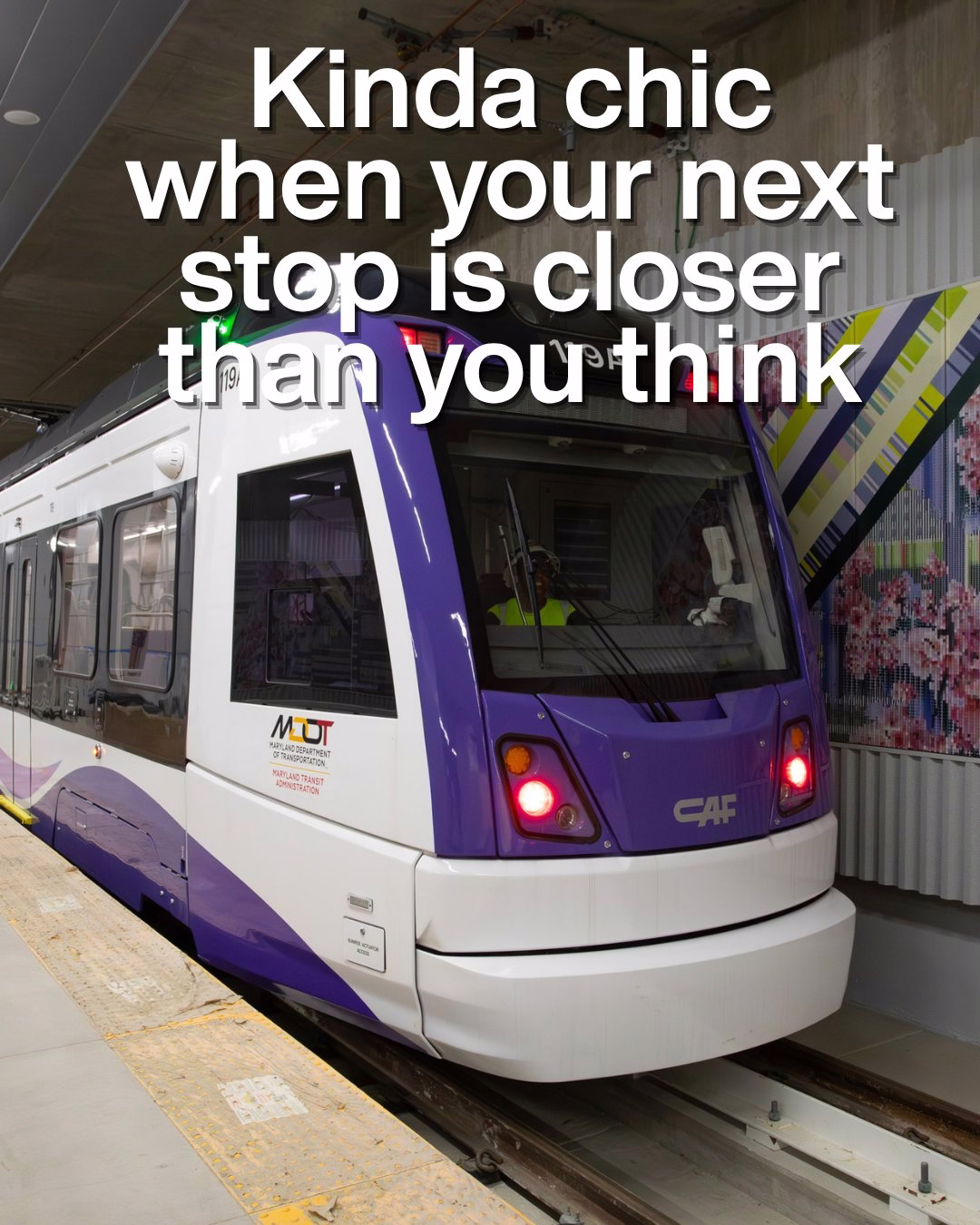 Modern purple and white light rail train stopped at an indoor station platform with colorful wall art nearby.